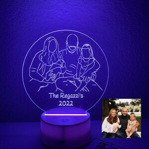 May include: A white acrylic night light with a custom outline of a family of four. The outline is illuminated in blue and the text "The Regazzi's 2022" is below the outline. A photo of the family is shown in the background.