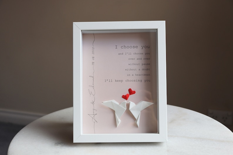May include: White frame with a pink background and a handwritten message that reads "Amy & Edmund 08.08.2020" and "I choose you and I'll choose you over and over without pause without a doubt in a heartbeat I'll keep choosing you". Two white origami doves are in the center of the frame with three red paper hearts above them.