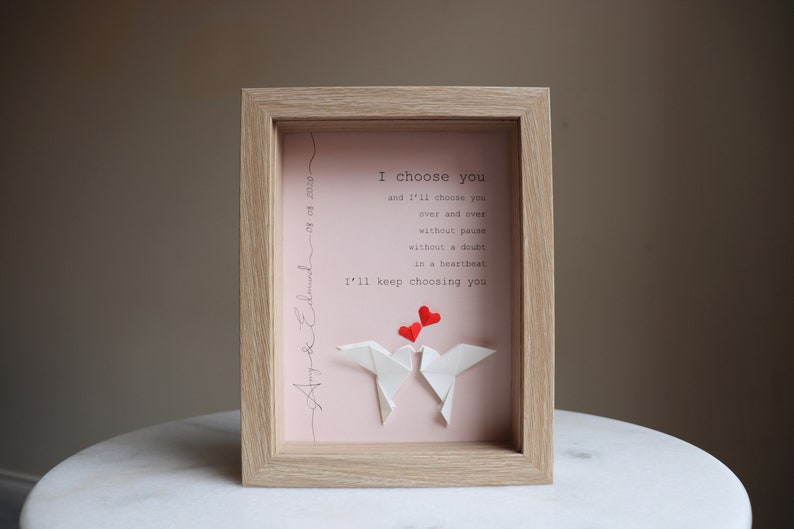 May include: A framed pink and white print with the text "I choose you and I'll choose you over and over without pause without a doubt in a heartbeat I'll keep choosing you". Two white origami doves are holding red hearts above them. The frame is light brown wood.