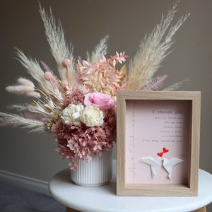 May include: A framed print with a pink background and the text "I choose you and I'll choose you over and over without pause without a doubt in a heartbeat I'll keep choosing you". Two red paper hearts are above two white paper origami birds.