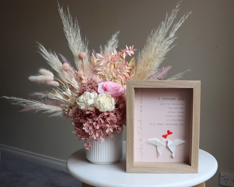 May include: A framed print with a pink background and the text "I choose you and I'll choose you over and over without pause without a doubt in a heartbeat I'll keep choosing you". Two red paper hearts are above two white paper origami birds.