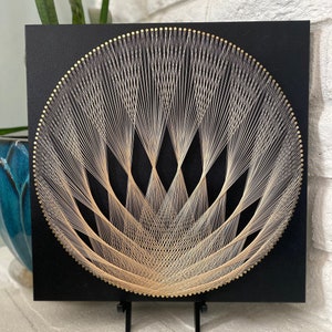 May include: A black square canvas with a gold string art design of a stylised flower. The flower is made of many thin strings that are stretched across the canvas and create a geometric pattern.