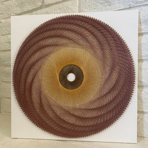 May include: A circular string art piece with a swirling pattern in shades of brown and pink. The center of the piece is a smaller circle with a white center.