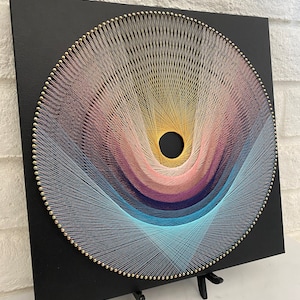 Op de afbeelding: Een cirkelvormig string art-stuk met een zwarte achtergrond. De string art is gemaakt met verschillende kleuren, waaronder roze, blauw en geel. De string art is gemonteerd op een zwart houten frame met een kleine zwarte ezel.
