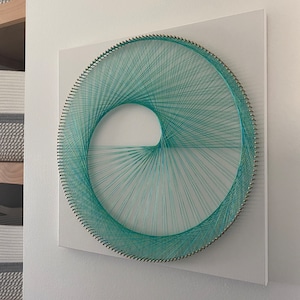 Puede incluir: Arte de hilo turquesa sobre un lienzo cuadrado blanco. El hilo está envuelto alrededor de pequeños clavos en un patrón circular, creando un diseño en espiral. La obra de arte está montada en una pared blanca, mostrando una estética moderna y minimalista.