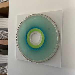 May include: A circular string art piece with a white background. The string art is made with blue, green, and white thread and is mounted on a white square board.