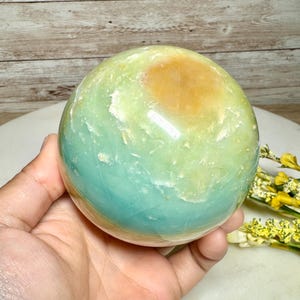 May include: A polished gemstone sphere with a blend of colors, including light green, blue-green, and a patch of golden-yellow. The sphere is held in a hand, showcasing its smooth, rounded form. The background features a wooden surface.