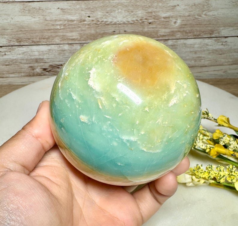 May include: A polished gemstone sphere with a blend of colors, including light green, blue-green, and a patch of golden-yellow. The sphere is held in a hand, showcasing its smooth, rounded form. The background features a wooden surface.