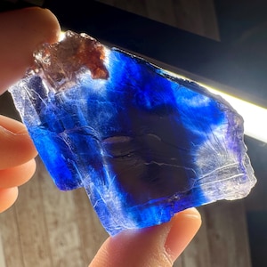Puede incluir: Un espécimen mineral translúcido de color azul, con áreas de índigo intenso y tonos más claros. La piedra, sostenida entre los dedos, muestra su estructura cristalina y variaciones naturales. La parte superior tiene una textura rugosa y marrón.
