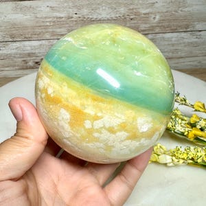 May include: A polished, spherical gemstone with bands of green, yellow, and white. The stone is held in a hand, showcasing its smooth, reflective surface. The natural stone has a unique color pattern and texture.