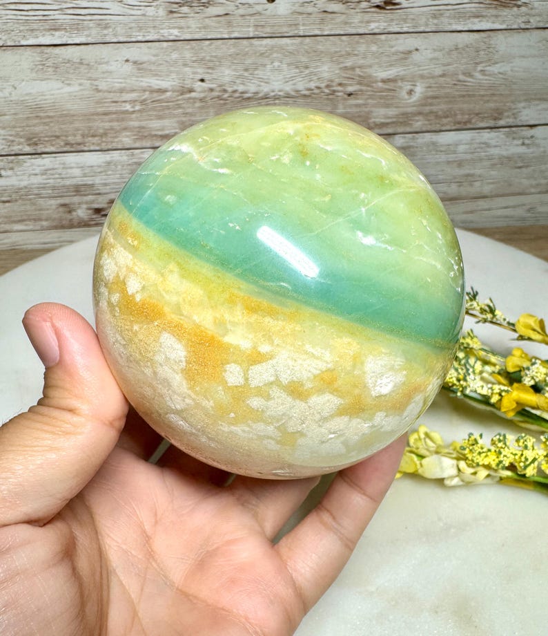 May include: A polished, spherical gemstone with bands of green, yellow, and white. The stone is held in a hand, showcasing its smooth, reflective surface. The natural stone has a unique color pattern and texture.
