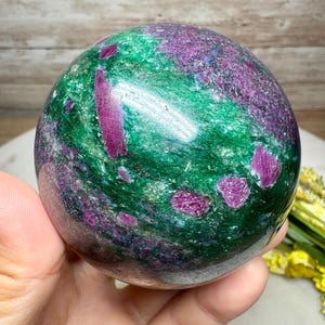 May include: A polished gemstone sphere, mainly green with striking magenta and purple inclusions. The sphere's surface reflects light, highlighting the natural patterns and textures. A decorative item for display.