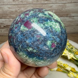 Peut inclure: Une sphère de pierre précieuse polie avec un mélange de couleurs. La sphère a une base bleu foncé et verte, avec des taches de rouge rubis. La pierre est lisse et ronde, avec une finition brillante. La sphère est tenue dans une main.