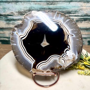 May include: A polished agate slice, showcasing concentric bands of black, white, and brown. The stone is mounted on a copper-colored metal stand. The natural stone features a central cavity with a unique crystalline formation.