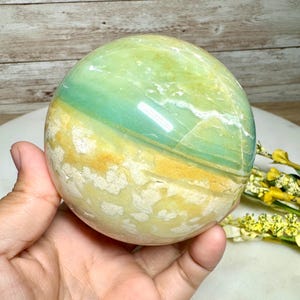 May include: A polished sphere of green, blue, and yellow stone. The orb is held in a hand, showcasing its smooth, reflective surface. The stone's colors blend in bands, creating a natural, earthy aesthetic. The sphere is a decorative item.