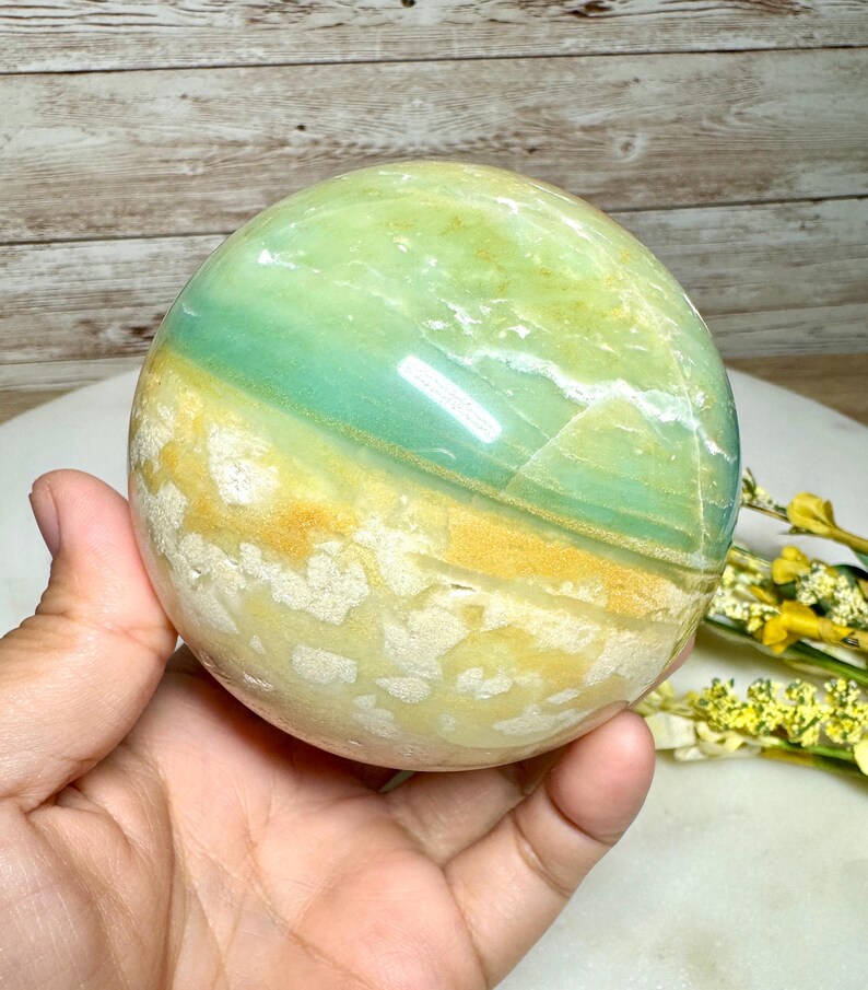 May include: A polished sphere of green, blue, and yellow stone. The orb is held in a hand, showcasing its smooth, reflective surface. The stone's colors blend in bands, creating a natural, earthy aesthetic. The sphere is a decorative item.