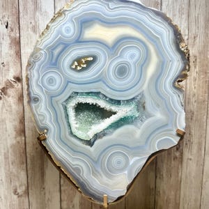 May include: A large, polished agate geode slice, showcasing concentric bands of gray, blue, and white. The center features a cavity filled with sparkling, light green crystals. The geode is mounted on a gold-toned metal stand, perfect for display.