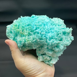 May include: A large, light blue and green crystal cluster with a rough, textured surface. The crystal is held in a person's hand.