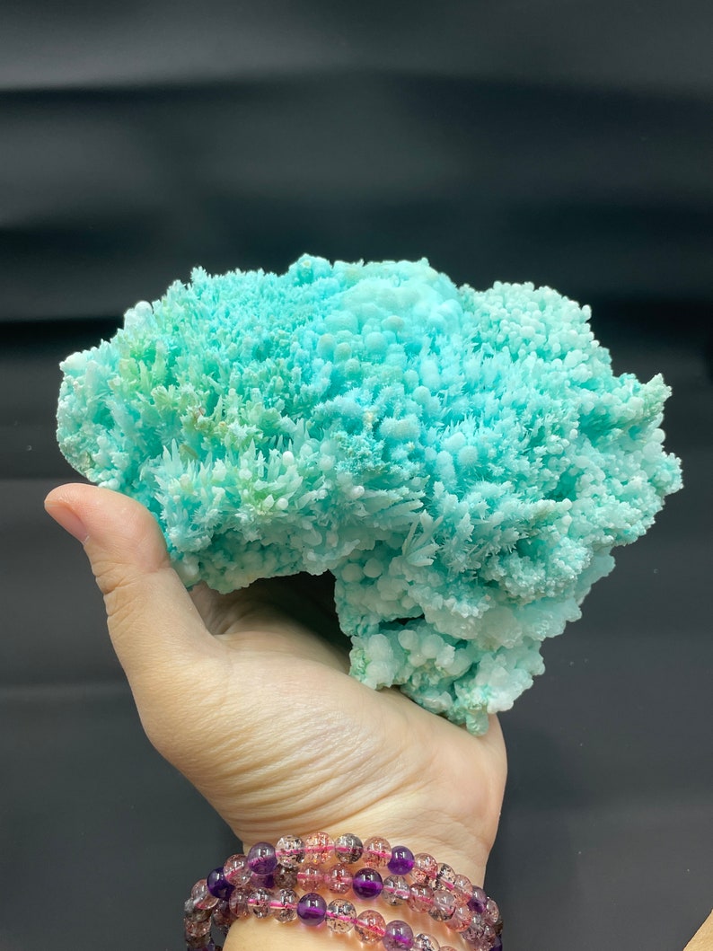 May include: A large, light blue and green crystal cluster with a rough, textured surface. The crystal is held in a person's hand.