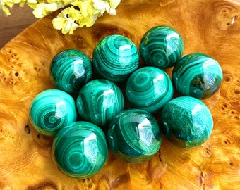 May include: A collection of ten green malachite spheres, each with unique swirling patterns, displayed in a wooden bowl.