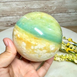 May include: A polished, spherical gemstone with bands of green, blue, and yellow. The orb is held in a hand, showcasing its smooth, reflective surface. The stone's colors create a natural, earthy aesthetic, suitable for home decor or crystal healing.