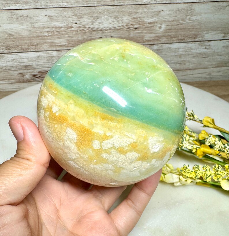 May include: A polished, spherical gemstone with bands of green, blue, and yellow. The orb is held in a hand, showcasing its smooth, reflective surface. The stone's colors create a natural, earthy aesthetic, suitable for home decor or crystal healing.
