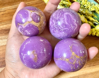 May include: Four purple and gold amethyst spheres, each with a unique pattern of swirling purple and gold veins. The spheres are smooth and polished, and they are displayed on a hand.