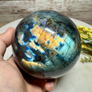 Peut inclure: Une sphère polie en labradorite, présentant un jeu de couleurs fascinant. L'orbite affiche des éclats irisés de bleu, d'or et de vert, créant un effet visuel captivant. La sphère est tenue dans une main, soulignant sa taille et sa forme.