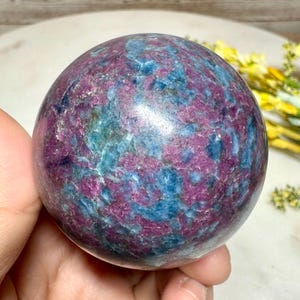 Könnte beinhalten: Eine polierte Kugel aus Rubin-Zoisit, gehalten in einer Hand. Der Stein weist ein gesprenkeltes Muster aus tiefem Magenta und Türkisblau auf, das einen auffälligen visuellen Kontrast erzeugt. Die Kugel ist glatt und abgerundet und zeigt die natürliche Schönheit des Minerals.