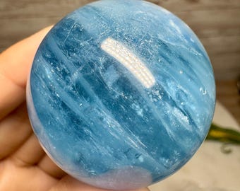 Puede incluir: Una gema pulida y esférica en tonos azul claro y medio. El orbe tiene una superficie lisa y reflectante con sutiles vetas internas. La gema se sostiene en una mano, mostrando su tamaño y color.