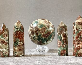 May include: Five polished green and red unakite crystal points and a sphere on a clear stand. The crystals are all different sizes and shapes.
