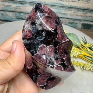 May include: A polished, freeform gemstone sculpture with a mix of deep burgundy, black, and white patterns. The stone has a smooth, curved shape and a glossy finish, showcasing its natural beauty. This decorative piece is ideal for home decor.