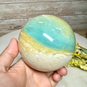 May include: A polished gemstone sphere with a gradient of colors. The sphere features bands of light blue, yellow, and white. The gemstone is held in a hand, with a wooden background.