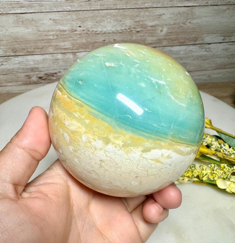 May include: A polished gemstone sphere with a gradient of colors. The sphere features bands of light blue, yellow, and white. The gemstone is held in a hand, with a wooden background.