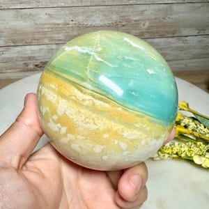 May include: A polished gemstone sphere with a blend of colors, including teal, yellow, and white. The sphere is held in a hand, showcasing its smooth, rounded form. The stone's surface displays natural patterns and variations.