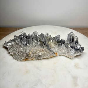 May include: A cluster of smoky quartz crystals, featuring a range of gray and clear tones. The crystals are densely packed, with sharp, pointed terminations. The base has a rough, textured appearance with hints of orange.