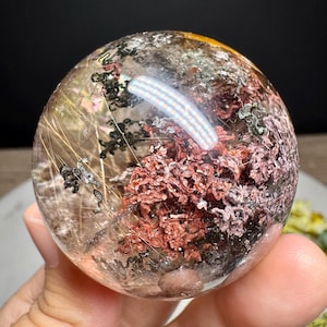 Pode incluir: Uma esfera de cristal de quartzo transparente contendo inclusões internas de formações minerais castanho-avermelhadas e verde-escuras, juntamente com finos fios dourados de rutilo em forma de agulha visíveis em toda a superfície da pedra polida.