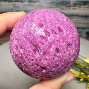 May include: A round, textured sphere of vibrant pink stone. The sphere is held in a hand, showcasing its color and surface details. The stone appears to be a natural mineral, possibly a decorative item or a collectible.