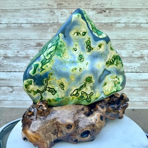 May include: A large, polished stone sculpture with a blue, green, and yellow pattern, mounted on a natural wood base. The stone has a smooth, glossy finish, and the wood base has a rustic, organic appearance. The sculpture is a decorative art piece.