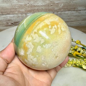 May include: A polished, spherical stone with a mix of colors. The stone features bands of green, yellow, and tan hues. The sphere is held in a hand, showcasing its smooth, rounded form. The stone has a natural, earthy appearance.