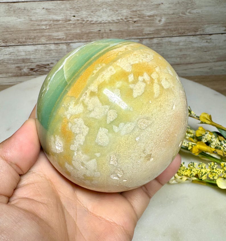 May include: A polished, spherical stone with a mix of colors. The stone features bands of green, yellow, and tan hues. The sphere is held in a hand, showcasing its smooth, rounded form. The stone has a natural, earthy appearance.