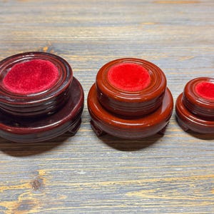 May include: Three wooden display stands in varying sizes. Each stand is a rich, dark brown with a glossy finish. The tops feature a circular, plush, red velvet insert. The stands are designed to showcase small objects.