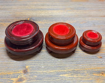 May include: Three wooden display stands in varying sizes. Each stand is a rich, dark brown with a glossy finish. The tops feature a circular, plush, red velvet insert. The stands are designed to showcase small objects.