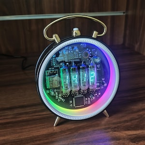 May include: A round digital clock with a black frame and gold-tone handle and feet. The clock face features four glowing green vacuum tubes displaying the time, set against an exposed circuit board with a vibrant, multi-colored LED ring light.
