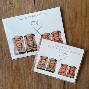 May include: Two wedding cards featuring a hiking boot theme. The top card reads "love is an adventure" with two hiking boots tied together with a heart. The bottom card reads "Nicole & Daniel congrats on your new adventure" with four hiking boots tied together with a heart.