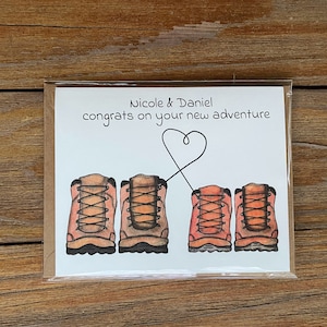 May include: A white card with the text "Nicole & Daniel congrats on your new adventure" and a drawing of four brown hiking boots with laces. Two of the boots are connected by a heart shape.