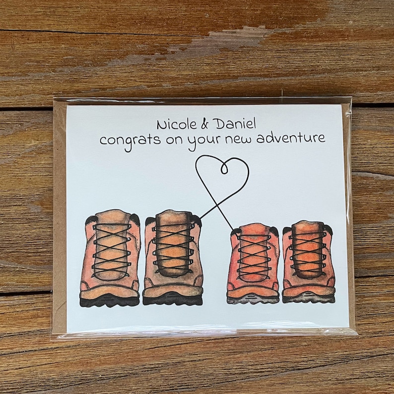 May include: A white card with the text "Nicole & Daniel congrats on your new adventure" and a drawing of four brown hiking boots with laces. Two of the boots are connected by a heart shape.