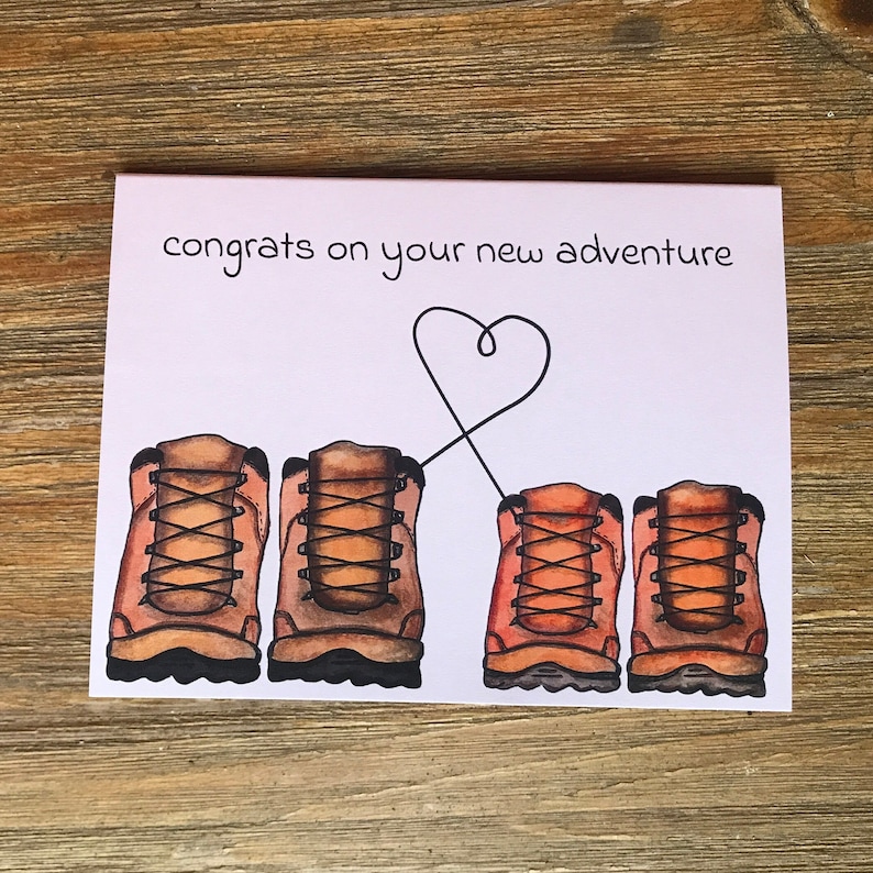 May include: A greeting card with the text "congrats on your new adventure" and a drawing of two pairs of brown hiking boots with a heart drawn between them.