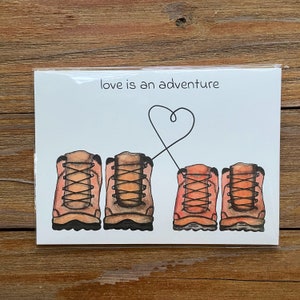 May include: A white card with the text "love is an adventure" and a drawing of four brown hiking boots with red laces. The boots are connected by a black line that forms a heart shape.