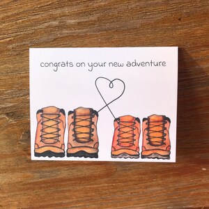 May include: A white card with the text "congrats on your new adventure" and a heart drawn with a black line. Four brown hiking boots are drawn below the text, with the two on the left connected by the heart line.